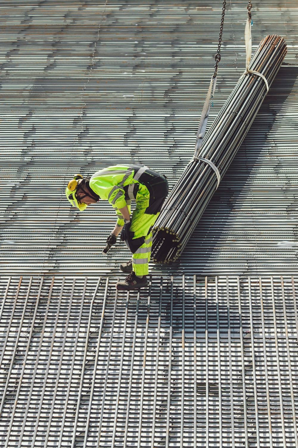 A worker in hi-vis guiding a crane-lifted bundle of rebar onto a reinforcement mesh floor, seen from above